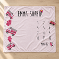 A personalized baby blanket laid flat on a light wood floor. The blanket is pale pink with black text reading 'EMMA SOPHIA' and a 12-month countdown grid labeled 'MONTHS'.