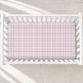 A top-down view of a white wooden crib containing a mattress fitted with a light pink and white houndstooth pattern sheet, set against a neutral cream textured background.