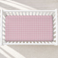 A pink and white plaid fitted sheet on a mattress inside a white wooden crib, shown from a top-down view on a light textured rug.