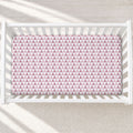 A top-down view of a white wooden crib with a pink and white geometric triangle patterned crib sheet, set against a light textured rug with soft window light shadows.