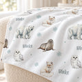 A white fleece throw blanket draped over a beige armchair, featuring watercolor illustrations of polar bears, seals, owls, foxes, and snowflakes, with the name 'Blake' printed in gray.