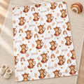 A white blanket with a repeating cartoon monkey print, rainbow motifs, and the name 'SKYLER' scattered across it, styled on a woven rug with small slippers and a wooden toy nearby.