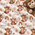 A close-up of a plush blanket with a repeating pattern of brown cartoon monkeys, orange leaves, small rainbows, and the name 'SKYLER' printed in brown. A brown plush monkey toy is partially visible in the top-right corner on a white background.