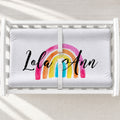 A white changing table with a personalized changing pad cover featuring a colorful watercolor rainbow and the personalized name 'Lola Ann' in black script, shown in a white crib with a safety strap.