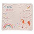 A personalized baby blanket featuring a white unicorn with orange and pink details, a pastel rainbow, scattered stars, and the name 'ELIZA grace' alongside a numbered calendar grid, all on a soft beige background.