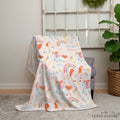 A pastel fleece throw blanket with a whimsical unicorn and rainbow print draped over a gray armchair in a styled room setting. The blanket features the names 'ELIZA' and 'GRACE' printed in blue and pink script, along with colorful unicorns, rainbows, and stars.