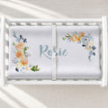 A white changing table with a personalized changing pad cover featuring a floral bumper sticker personalized with the name 'Rosie' in light blue script. The floral design features peach, yellow, and blue watercolor flowers with green leaves.