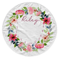 A round white baby blanket featuring a colorful watercolor floral wreath with the name 'Riley' in script and numbers 1 through 12 arranged around the border for monthly milestone tracking.