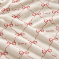 Close-up of a soft cream-colored fabric with a repeating pattern of dusty pink bow outlines and the word 'Avery' embroidered in matching pink script.