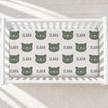 A fitted crib mattress with a repeating pattern of dark green cat faces and the name 'ELARA' in matching green text, shown inside a white wooden crib on a light neutral floor.