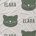 A close-up of a white fabric with a repeating pattern of dark green cat faces and the name 'ELARA' in dark green text.