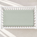 A top-down view of a white wooden baby crib with a sage green and white gingham checkered mattress cover, set against a light beige textured rug with soft diagonal shadows.
