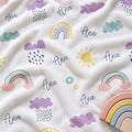 A close-up of a white baby blanket or cloth printed with pastel rainbows, clouds, suns, and the name 'Ana' in script, with a wooden rainbow toy partially visible at the bottom right.