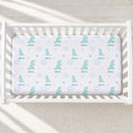 A white crib mattress fitted sheet with a repeating pattern of light blue dinosaurs, hearts, stars, and the name 'Ailana' printed in blue, shown inside a white wooden crib.