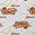 A close-up of a white fabric with a repeating watercolor-style print featuring brown sloths resting on branches, yellow crescent moons, small gold stars, and the name 'Sienna' in brown script.