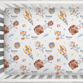 A fitted crib mattress cover with a repeating space-themed print, featuring cartoon astronauts, rockets, planets, stars, and the name 'Rowan QUINN' in blue and orange text, shown inside a white wooden crib.