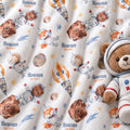A close-up of a plush blanket or fabric featuring a repeating astronaut teddy bear print with the name 'Rowan Quinn' and small planets/stars. A plush teddy bear wearing a space suit is placed on the right side as a prop.