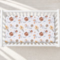 A white crib mattress fitted sheet with a playful space-themed print featuring cartoon astronauts, rockets, planets, and the name 'Rowan' printed repeatedly, shown inside a white wooden crib.