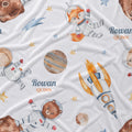 A white fabric blanket or throw printed with a whimsical space-themed pattern featuring watercolor-style astronauts, a fox, a bear, a rocket, planets, stars, and the personalized name 'Rowan Quinn'.