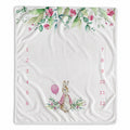 A baby milestone blanket featuring a watercolor floral border in green and pink at the top, a small bunny illustration with a pink balloon near the bottom center, and red printed numbers 1 through 12 arranged in two columns on either side.