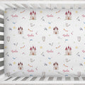 A fitted crib sheet with a white background and a repeating pattern of fairytale castles, bows, arrows, flags, and the word 'Gala' in pink script, shown inside a white wooden crib.