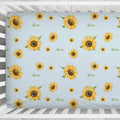 A personalized baby crib mattress cover featuring a light blue background with a repeating pattern of yellow sunflowers and the name 'Alexa' printed in green script. The cover is shown fitted inside a white wooden crib.
