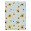 A square fabric item, likely a small blanket or towel, featuring a repeating pattern of yellow sunflowers and the name 'Alexa' in green cursive on a light blue background.