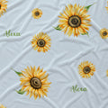 A light blue fabric with a repeating pattern of yellow sunflowers and the name 'Alexa' printed in green script.