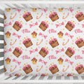 A crib mattress fitted sheet with a repeating pattern of watercolor cupcakes, pink lollipops, candy canes, and the name 'Ellie' in pink script, shown on a white wooden crib.