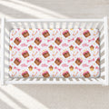 A fitted crib sheet with a repeating pattern of cupcakes, pink bows, candy canes, and the name 'Ellie' in script, shown inside a white wooden crib on a light floor.