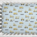 A personalized baby crib mattress cover with a light blue background, featuring a repeating pattern of yellow construction vehicles and the name 'DANIEL' in dark gray. The cover is shown fitted inside a white wooden crib.