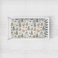 A baby crib with white slats, fitted with a printed sheet featuring teddy bears, boats, and the name 'Oliver' in a vintage illustration style, set against a light whitewashed wood background.