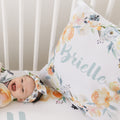 A baby lying on a white crib mattress wearing a matching floral headband, next to a large floral pillow printed with the name Brielle in script.
