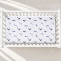 A white crib mattress fitted sheet with a navy blue whale print and the name 'James' repeated, shown inside a white wooden crib on a light neutral floor.
