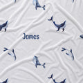 A white fabric item, likely a baby blanket or swaddle, featuring a repeating pattern of watercolor blue and gray whales and the name 'James' printed in navy blue text in the center.