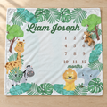 A personalized baby blanket laid flat on a light wood floor. The white blanket features a colorful jungle-themed border with green leaves, cartoon animals including a giraffe, zebra, lion, elephant, and monkey, and the text 'Liam Joseph' at the top plus a 12-month calendar grid in the center.