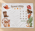 A personalized fleece blanket with a western theme, laid flat on a light wood floor. The blanket features the name 'Scout Ellis' at the top, a 12-month countdown grid, and various western illustrations including a cowboy hat, boots, cactus, and money bag.