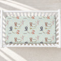 A top-down view of a white wooden crib with a custom printed mattress protector featuring woodland animal illustrations and the name 'Charlie' on a pale mint green background.