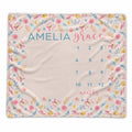 A personalized baby blanket with a floral border and a central baby name and calendar grid. The blanket features the name 'AMELIA grace' in blue and pink lettering, with numbers 1 through 12 arranged for tracking months.