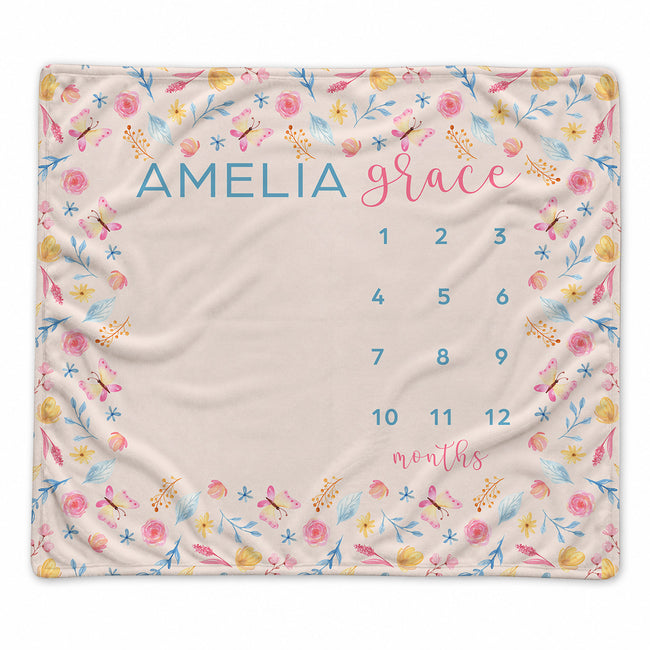 A personalized baby blanket with a floral border and a central baby name and calendar grid. The blanket features the name 'AMELIA grace' in blue and pink lettering, with numbers 1 through 12 arranged for tracking months.