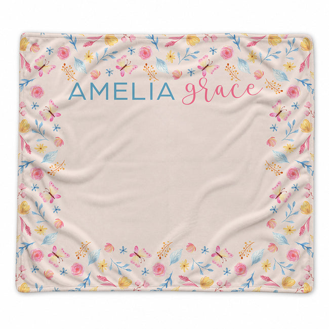 A square personalized blanket with a cream background, decorated with a colorful floral and butterfly border and the names 'AMELIA' and 'grace' printed at the top center.