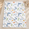 A square white baby blanket with an underwater theme, featuring blue dolphins, orange fish, green seaweed, and the word 'SILAS' repeated, laid flat on a beige carpet next to a teddy bear.