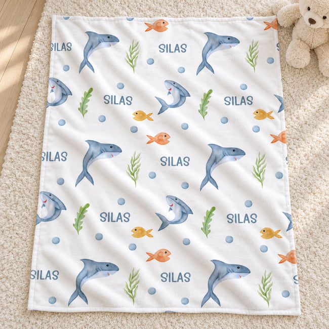 A square white baby blanket with an underwater theme, featuring blue dolphins, orange fish, green seaweed, and the word 'SILAS' repeated, laid flat on a beige carpet next to a teddy bear.