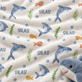 A close-up of a plush white fabric printed with blue sharks, small orange fish, green seaweed, blue dots, and the word 'SILAS' repeated. A blue-and-white plush shark toy is partially visible in the lower right corner.