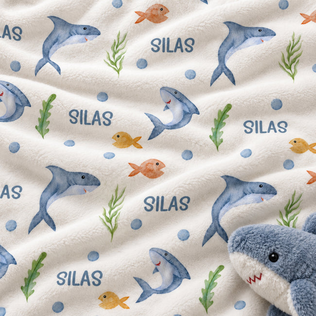 A close-up of a plush white fabric printed with blue sharks, small orange fish, green seaweed, blue dots, and the word 'SILAS' repeated. A blue-and-white plush shark toy is partially visible in the lower right corner.