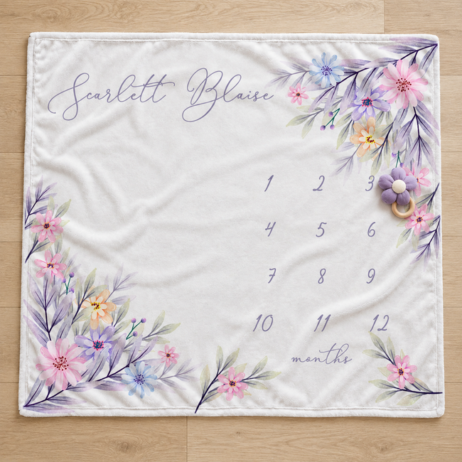 A square white baby blanket laid flat on a light wood floor, printed with the name 'Scarlett Blaise' in script at the top left, a floral border in pastel pink, purple, blue, and yellow around the edges, and a numbered monthly calendar from 1 to 12 with the word 'months' near the bottom.