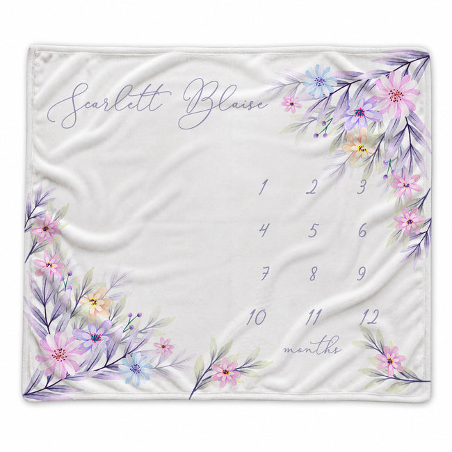 A personalized baby blanket featuring a watercolor floral design in pink, purple, and blue around the border, with the name 'Scarlett Blaise' in script and a numbered 1-12 month grid on a white background.