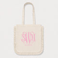 An off-white canvas tote bag with fringed edges and long handles, featuring a candy pink monogram 'SWM' and the name 'SLOANE' printed on the front, set against a light gray background.