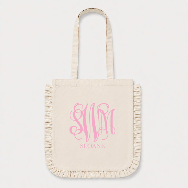 An off-white canvas tote bag with fringed edges and long handles, featuring a candy pink monogram 'SWM' and the name 'SLOANE' printed on the front, set against a light gray background.