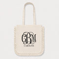 An off-white canvas tote bag with fringed edges and long handles, featuring a black monogram 'CBM' and the name 'CAROLINE' printed on the front, set against a light gray background.